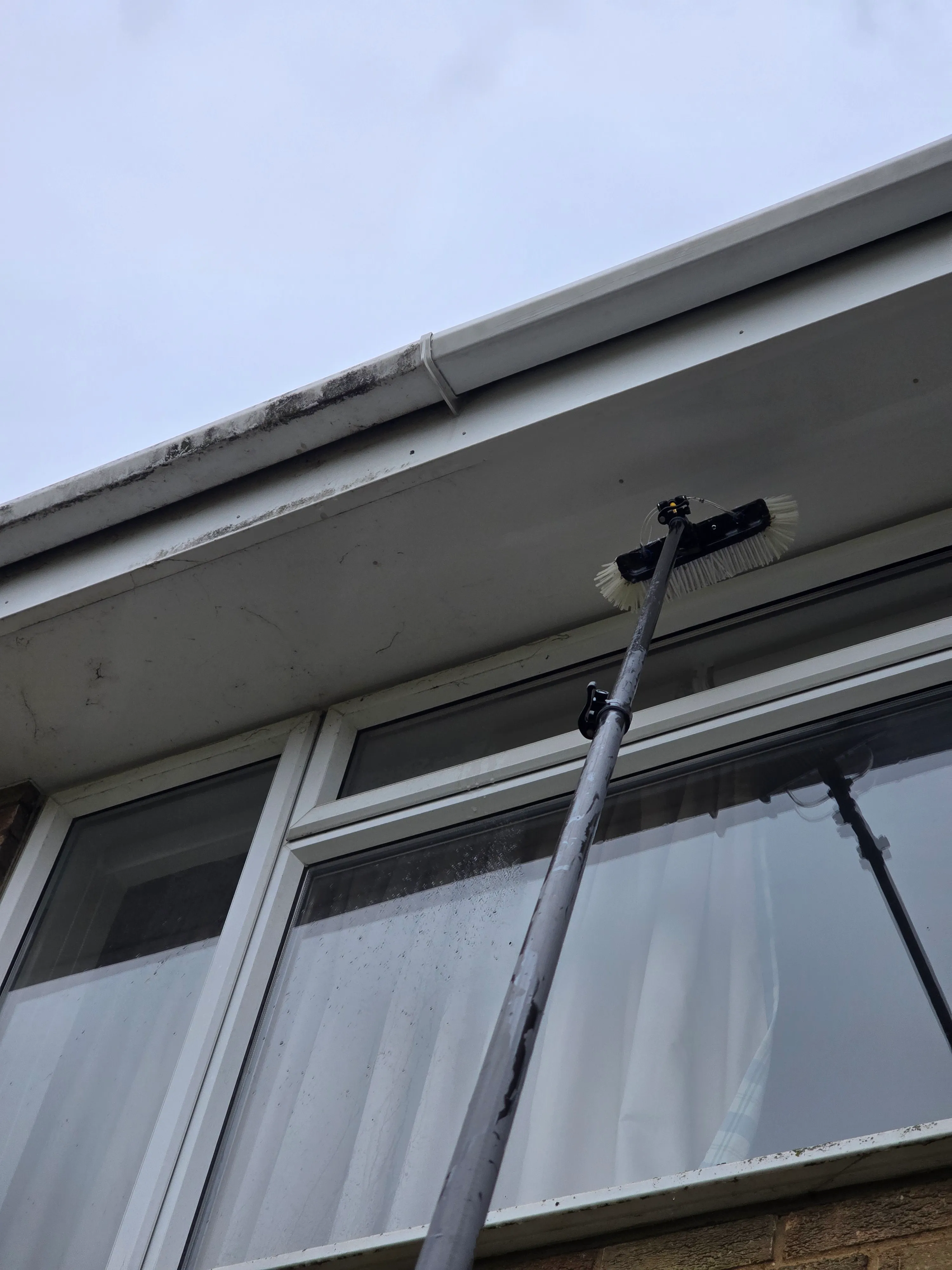 A soffit and fascia part-way through cleaning. The right-hand stretch is clean white; the left is still grey and streaked with green algae.