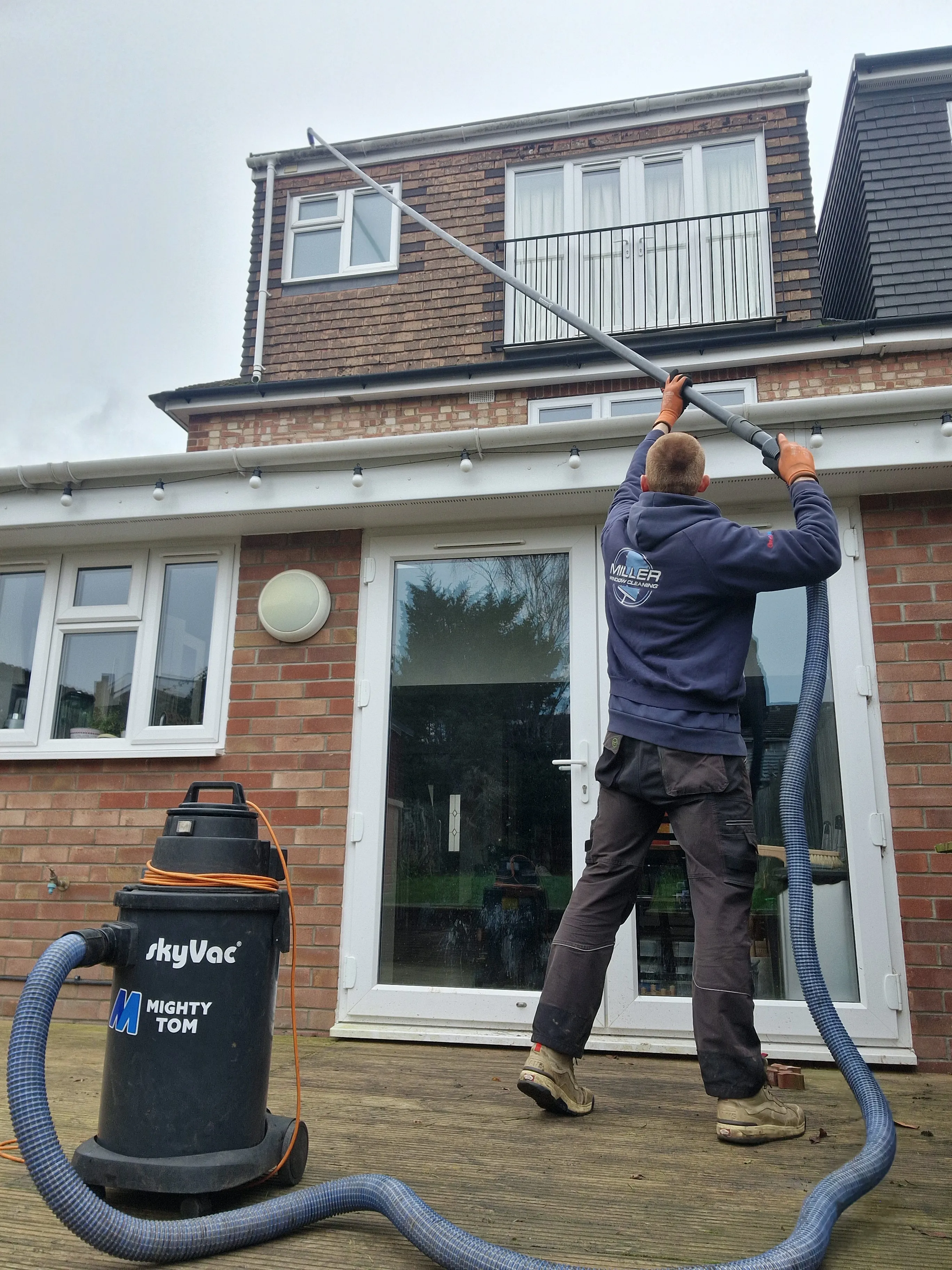 A gutter vacuum standing on the decking with its hose running up a long pole into the gutter of a house, operated from the ground by a cleaner in a Miller Window Cleaning hoodie.