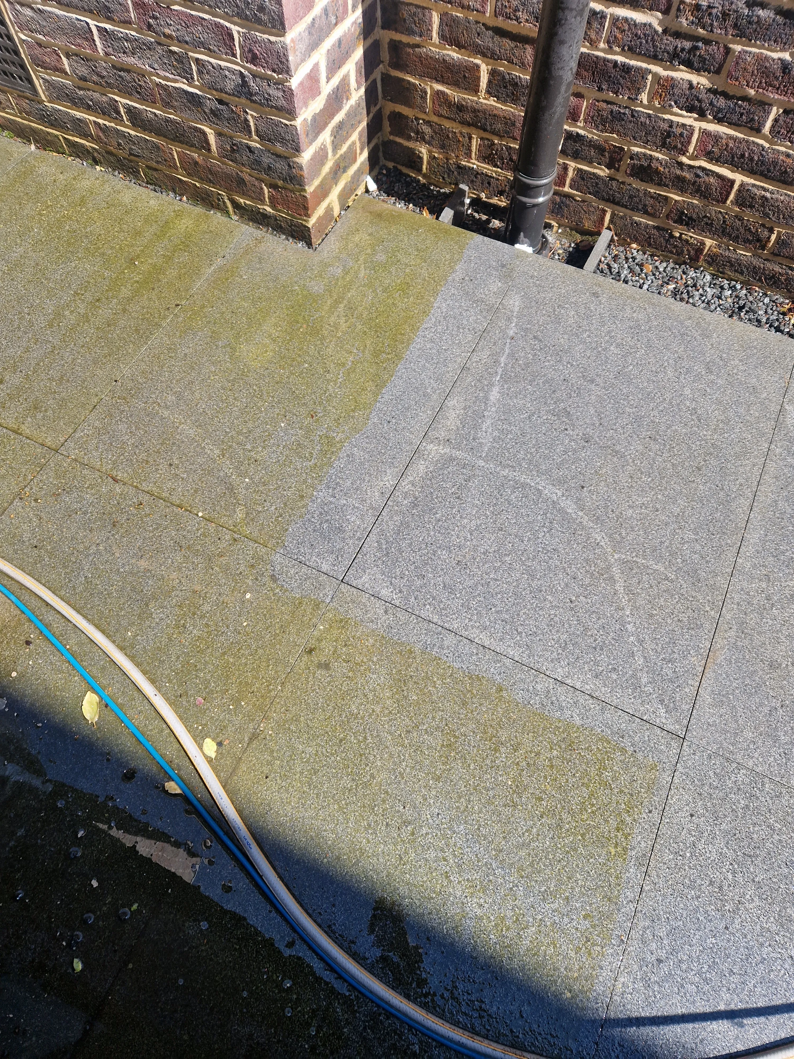 A patio part-way through pressure washing. A band of slabs has been cleaned back to pale grey stone, while the slabs around it are still green with algae.