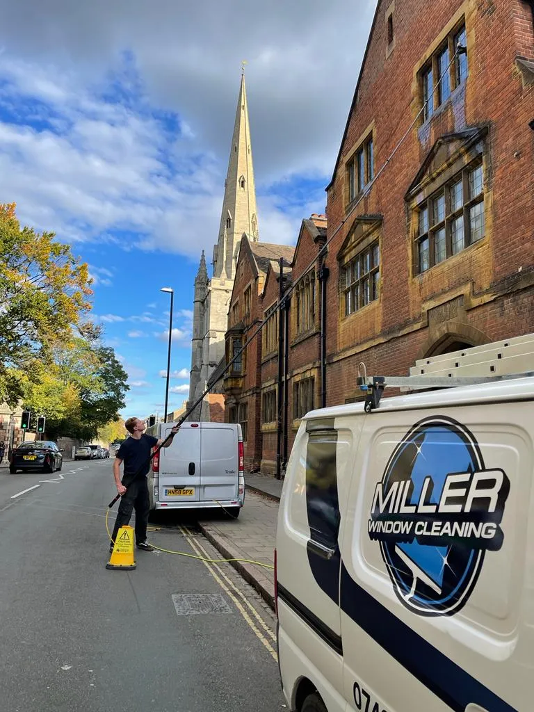 A Miller Window Cleaning van parked on a Cambridge street while a cleaner uses a water-fed pole on the upper windows of a brick building, with a church spire behind.